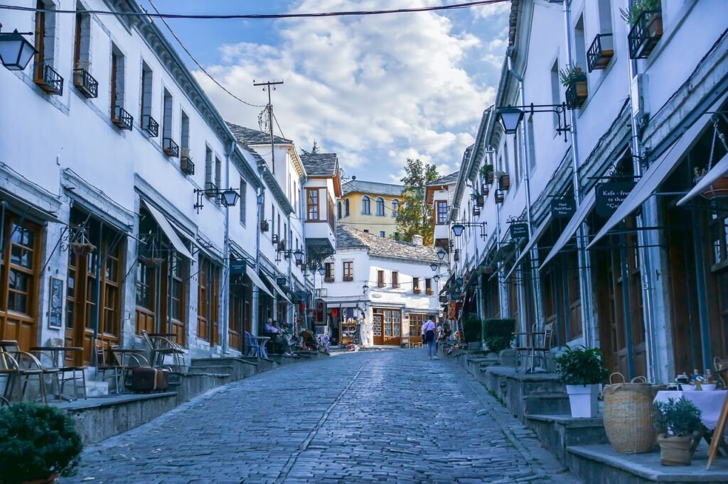 Vibrant street in Gjirokaster featuring cafes, grey stoned houses, and small shops with people walking.