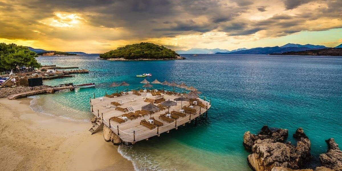 Sunset view of Ksamil Island with golden light illuminating azure waters and sandy shore.