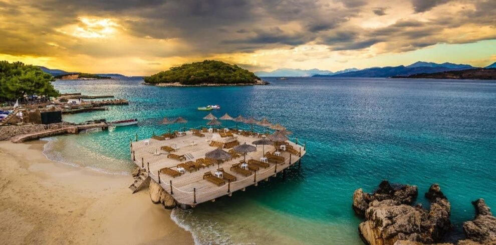 Sunset view of Ksamil Island with golden light illuminating azure waters and sandy shore.