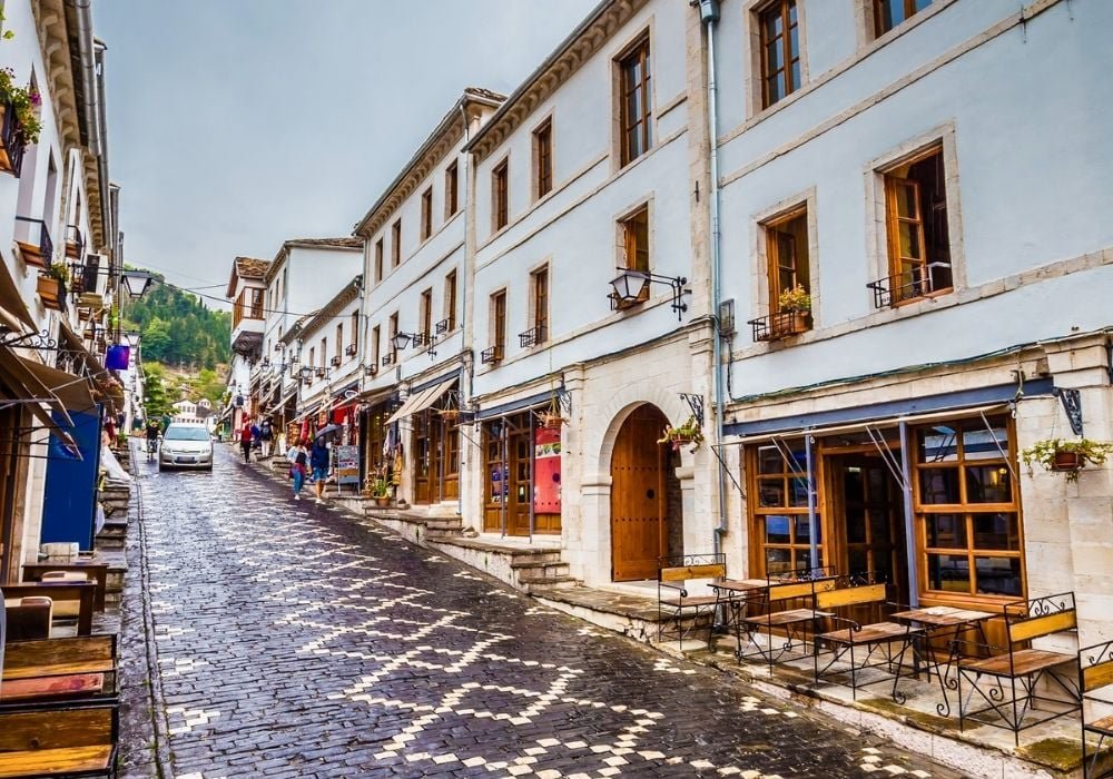 Vibrant street in Gjirokaster featuring cafes, wooden-window houses, and small shops with people walking.